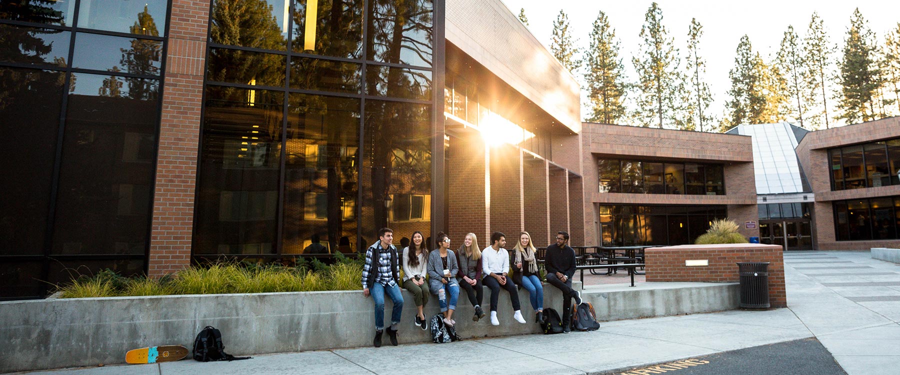 Students sit on a wall outside a large, modern academic building. Setting sunlight hits the windows behind them.