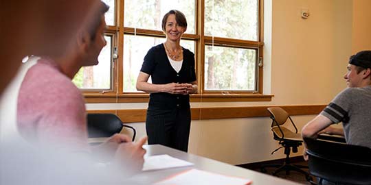 A professor stands in a classroom near a large window, smiling and listening to a student.