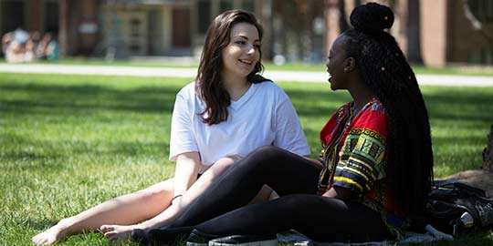 Two students of different ethnicities sit together, conversing, on the manicured lawn outside an academic building.