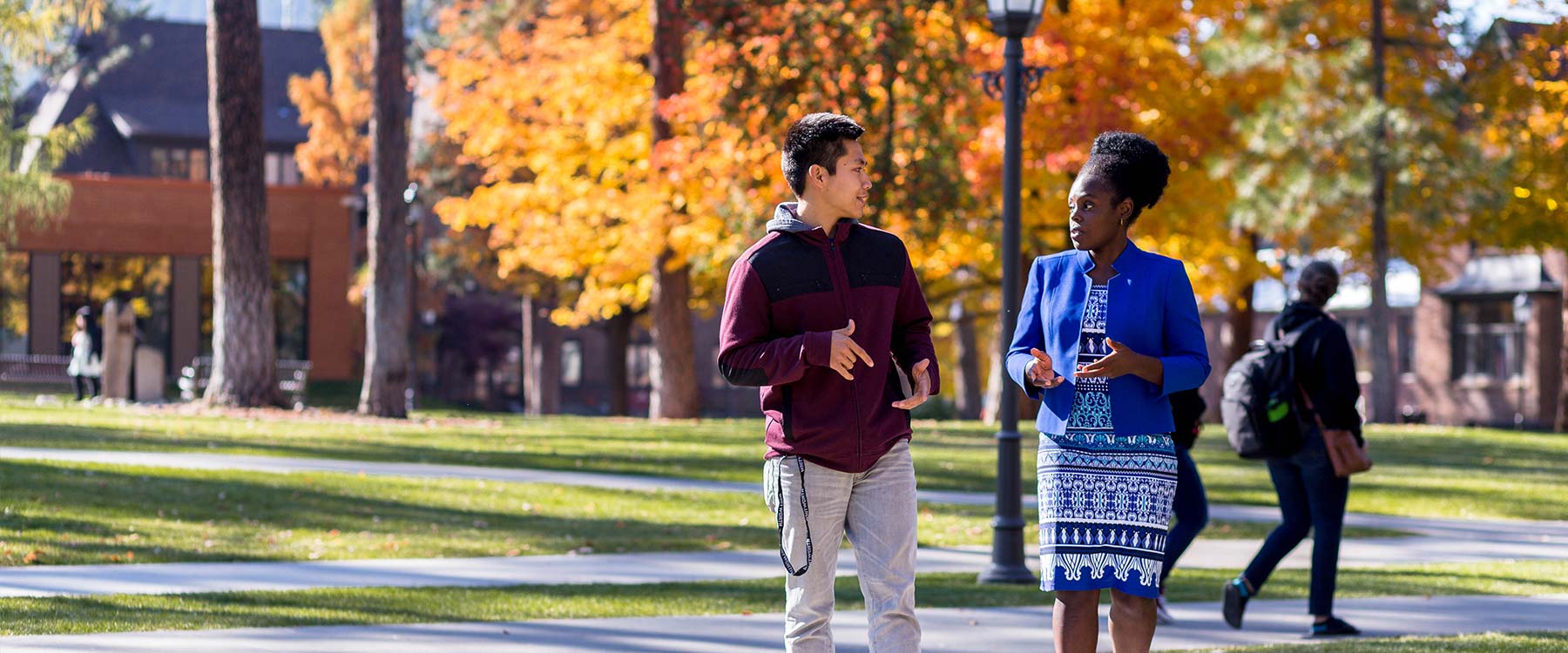 A professor talks with a student while walking on campus on a sunny autumn day.