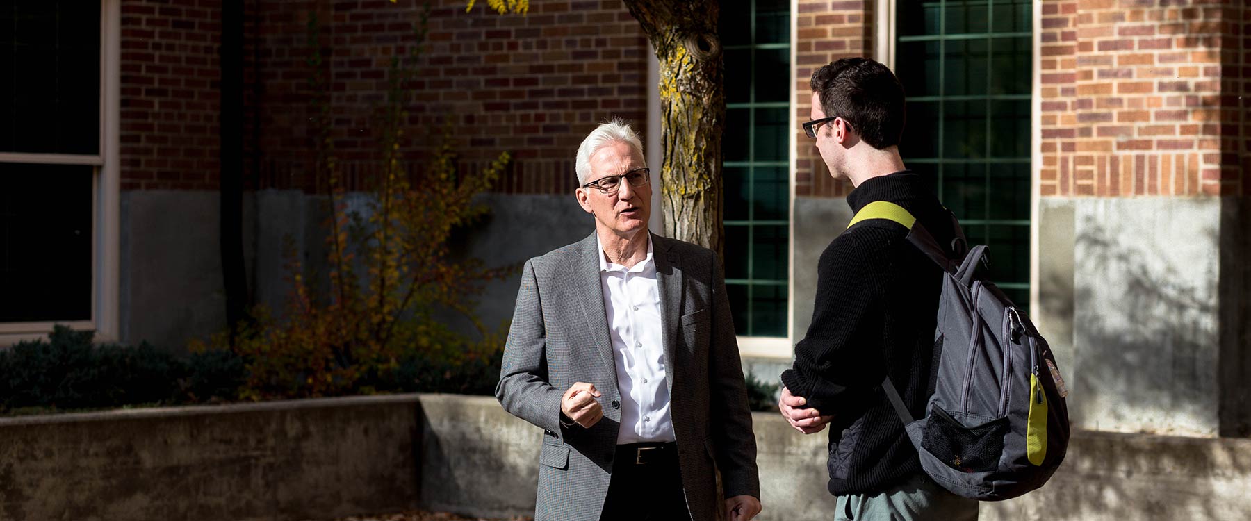 A professor stands and speaks with a student outside a large academic building in the sun.