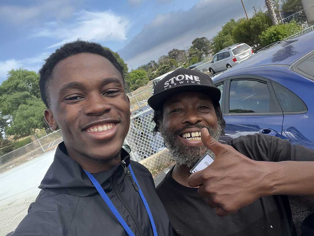 Two people face the camera for a selfie. Both have big smiles, and one gestures with a thumbs up. Behind them is a blue sky with swirling clouds.