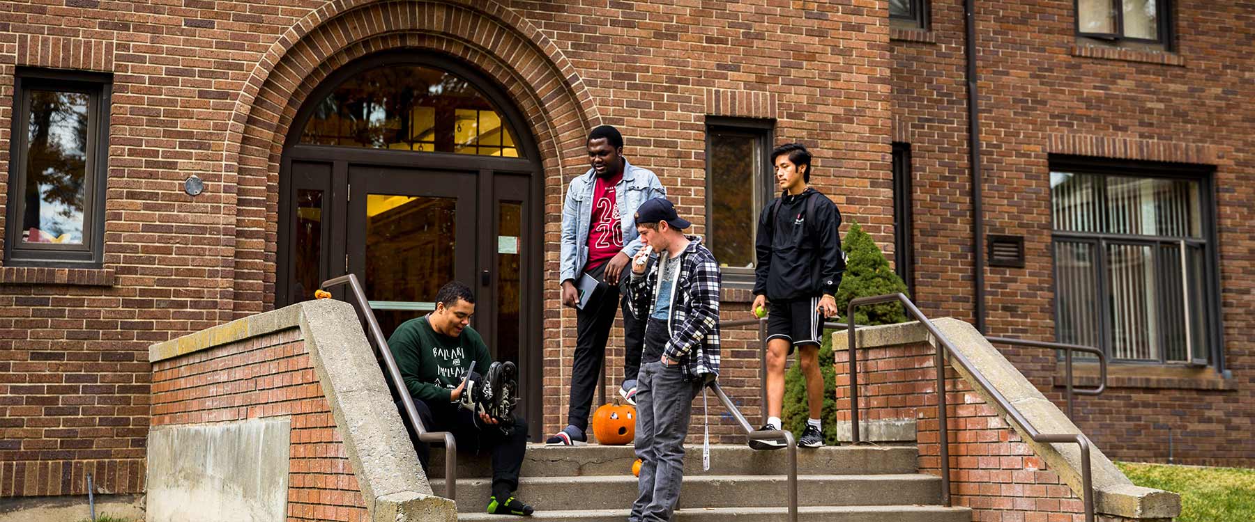Four students gather on the steps outside of McMillan Hall. They watch as one student puts on roller blades.