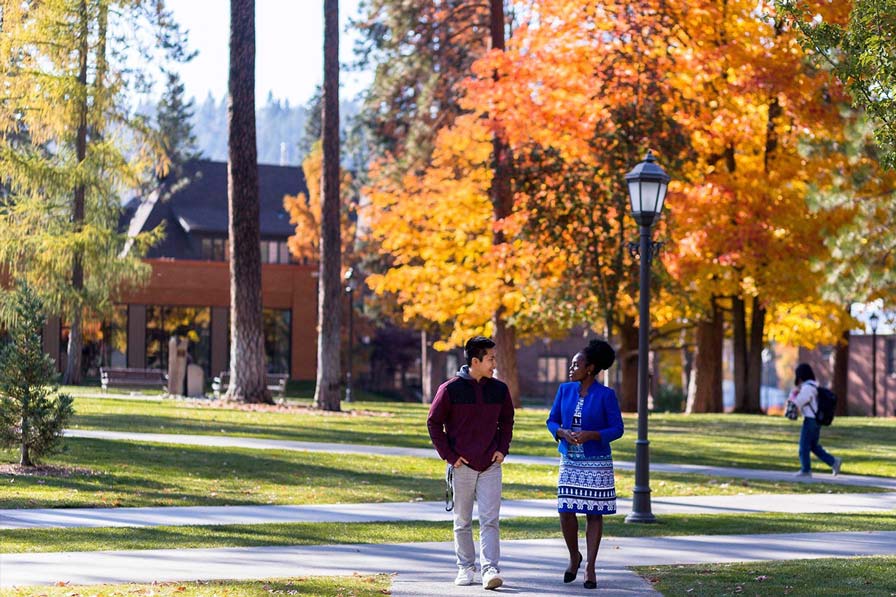 A student and professor walk on a tree-lined path with vibrant autumn foliage.