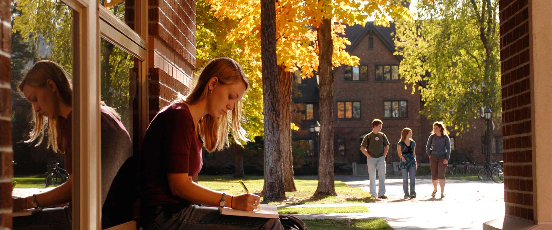 A student sits on a ledge outside an academic building and highlights a textbook. Three students walk toward the building.