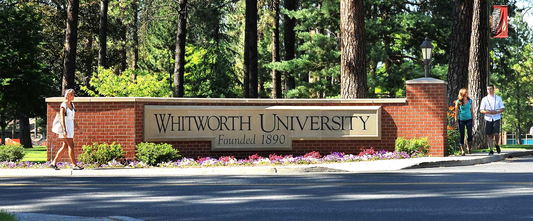 Students walk on the sidewalk on either side of a brick Whitworth campus entrance sign.