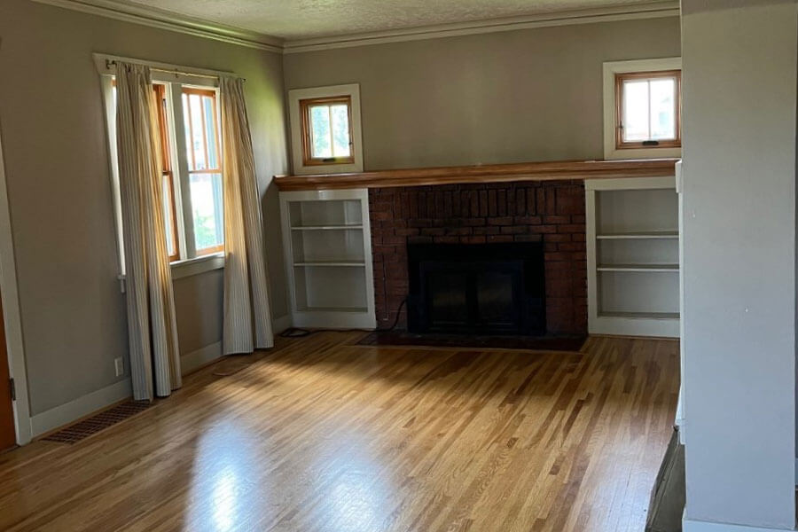 Empty living room with hardwood floors, fireplace, built-in shelves and windows with curtains.