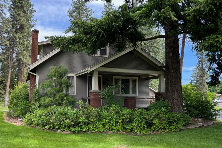 A classic gray house with brick accents and chimney, surrounded by lush greenery and tall trees.