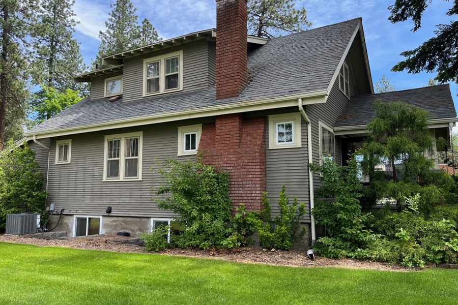 Side view of a gray, two-story house with a red brick chimney, surrounded by trees and greenery.