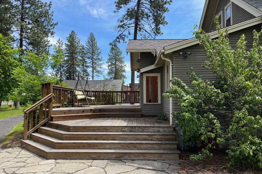 Large wooden deck leading from a stone patio to a craftsman style house, surrounded by pine trees.