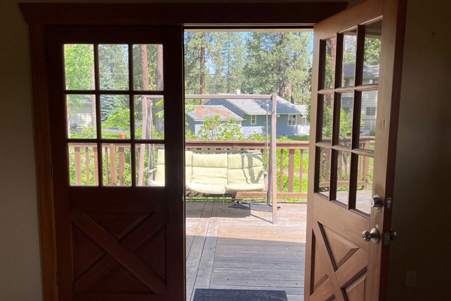 Open double doors lead to a wooden deck with a swinging bench, with foliage and houses in the distance.