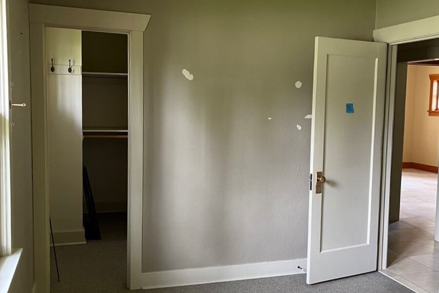 Empty bedroom with gray walls and carpet, an open closet with shelves and hooks, and a doorway out to the main living area.