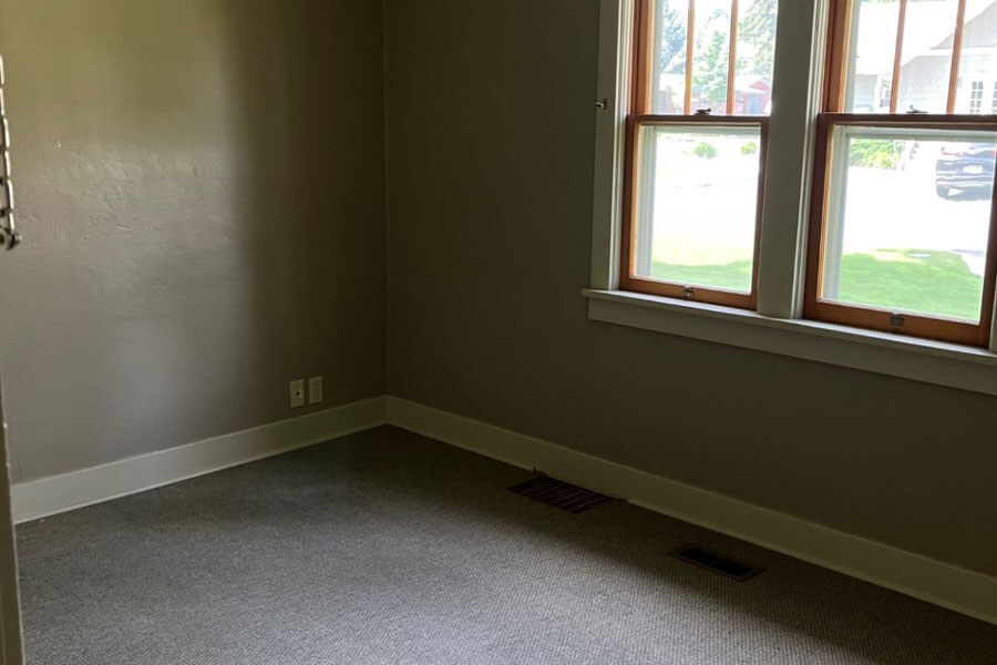 Empty bedroom with gray walls, a carpeted floor, and large wood-framed windows looking over the lawn.