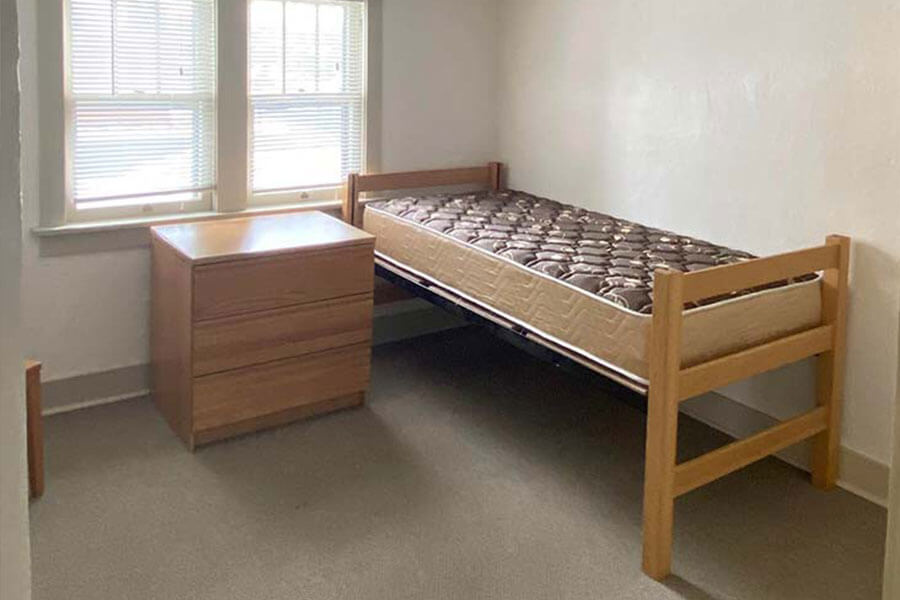 Minimalist room with a single bed, wooden dresser, and window with blinds.