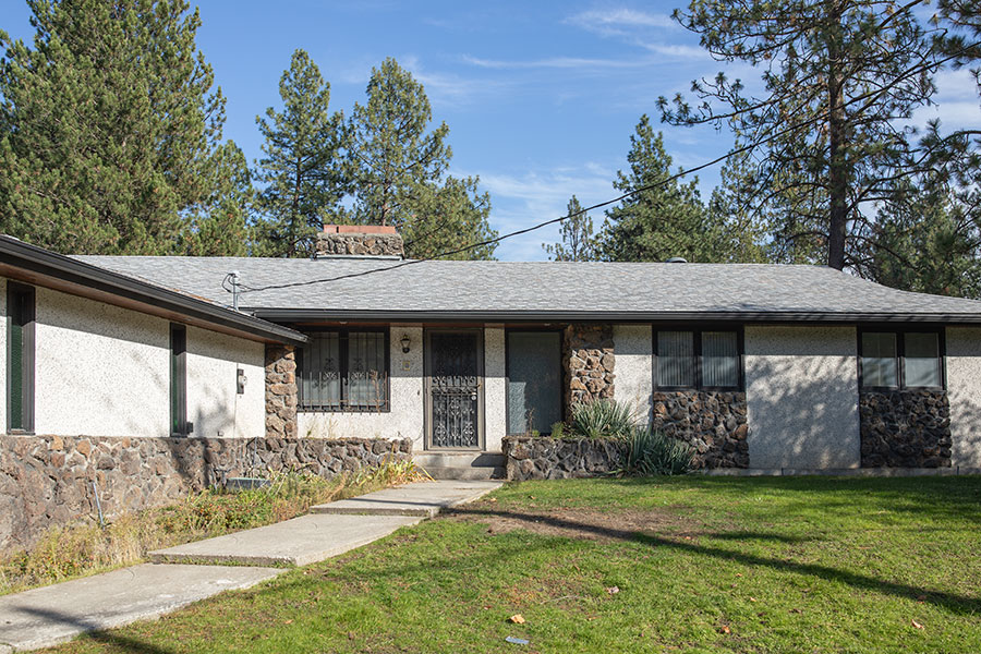 Single-story house with stone facade, surrounded by trees and a grassy lawn under a blue sky.