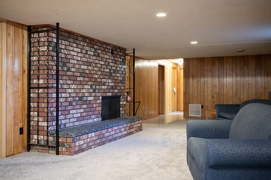 A cozy basement rec room with wood paneling, a brick fireplace and lounge furniture.