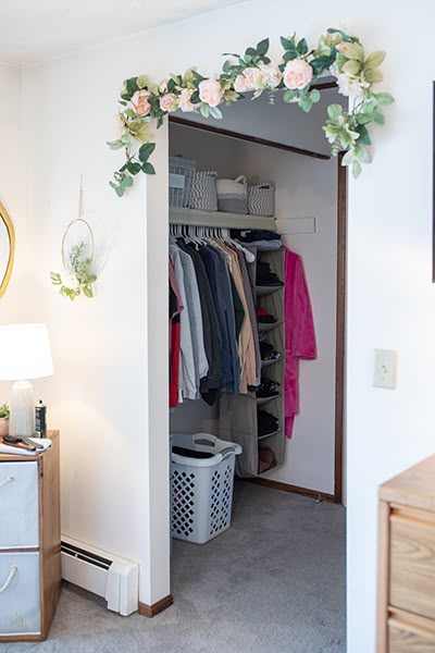 Open closet with clothes, a laundry basket, and floral arch decoration above the doorway in a cozy room.