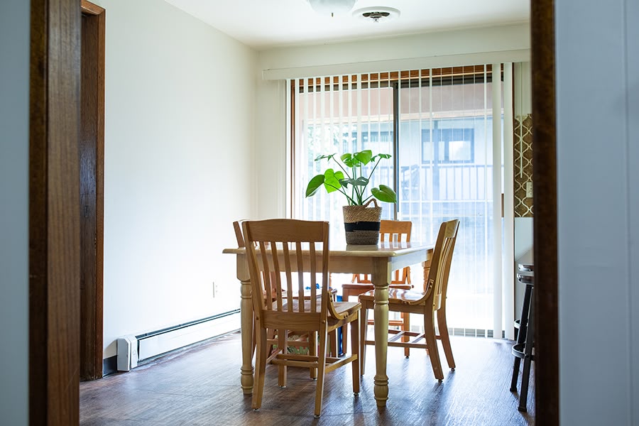 A sunlit kitchen nook with a wooden table and four chairs near a sliding glass door. A potted plant sits on the table.