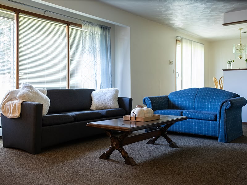 Living room with two sofas, a wooden coffee table, and large windows with blinds and curtains.