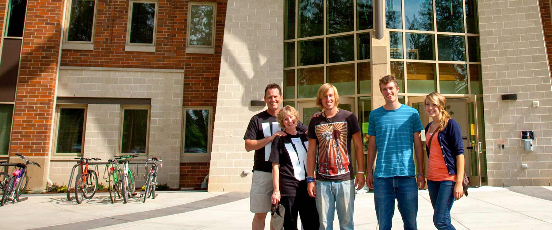 Two parents and three grown kids pose, smiling, outside a campus residence hall.