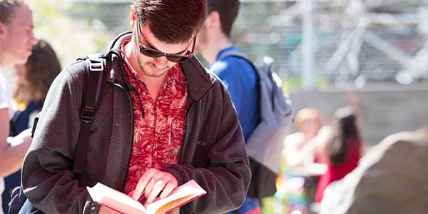 A student wearing sunglasses reads an open book in a crowded group setting on a sunny day.