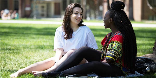 Two students of different ethnicities sit together conversing on the manicured lawn outside an academic building.