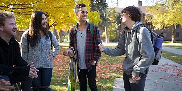Four students laugh and converse outside an academic building on a fall day.