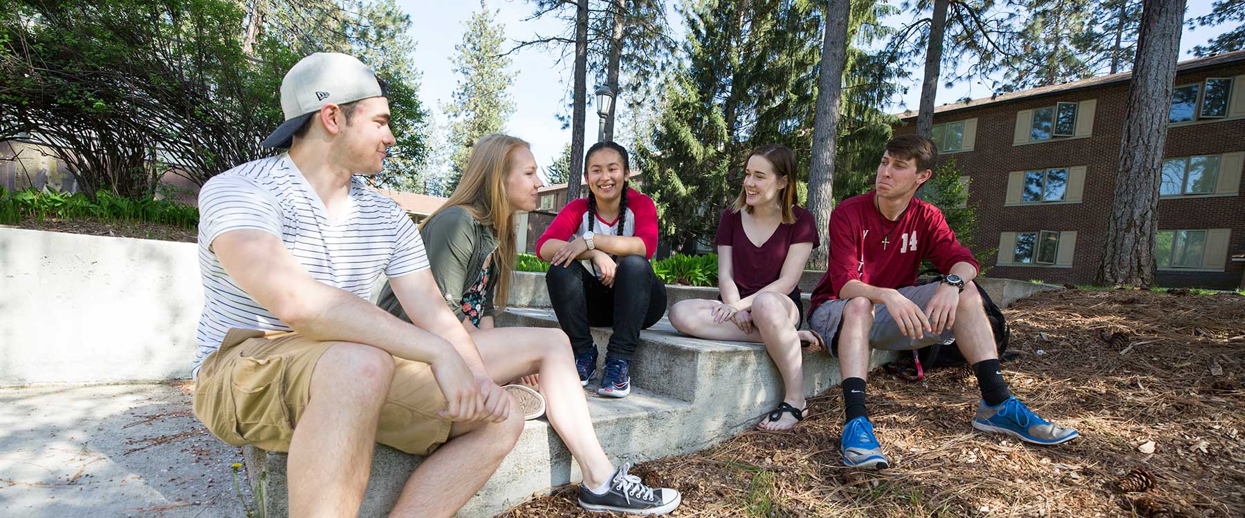 Five students sitting on steps outdoors, smiling and chatting, with trees and a building in the background.