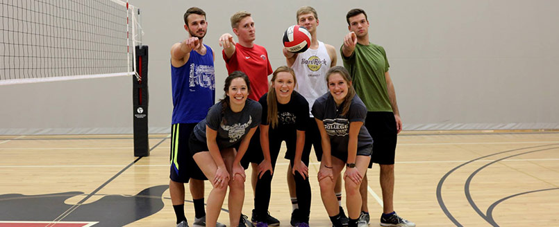 Student-athletes pose together on a court, with one holding a volleyball and several pointing toward the camera.