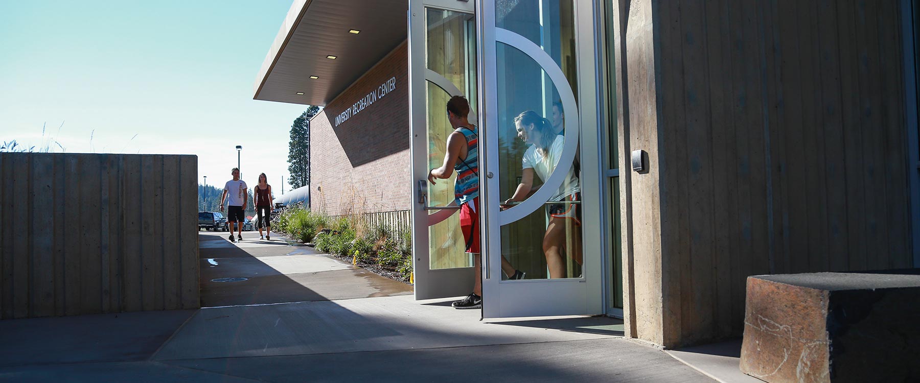 Two students push open the doors of the U-Rec, a modern building with glass doors, as they leave.