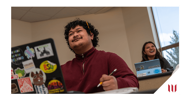 Two students sit at desks in a classroom. Both have laptops open and are smiling.