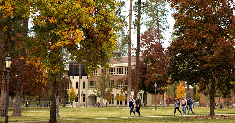 Students walk in groups between stately, brick academic buildings on a scenic fall day.