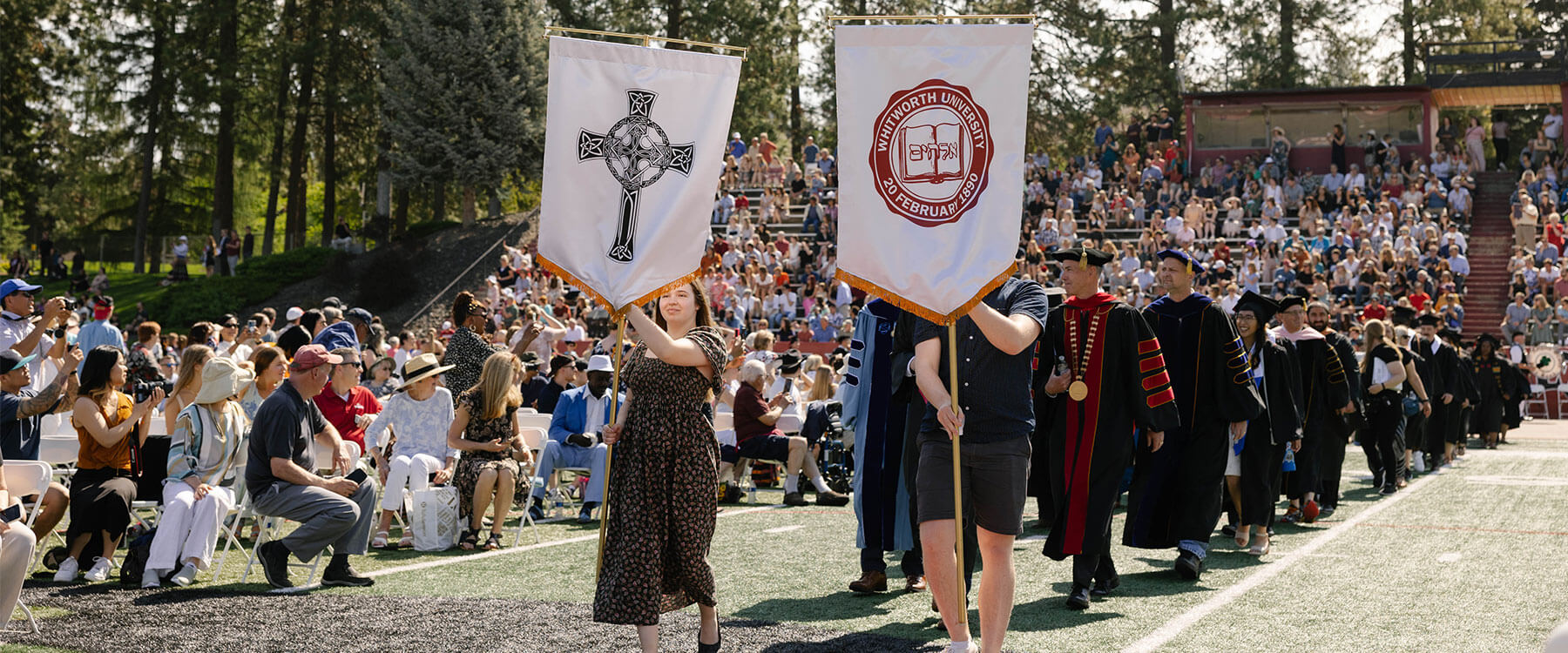 A large group of people in academic regalia participate in a ceremonial procession, holding banners with symbols.