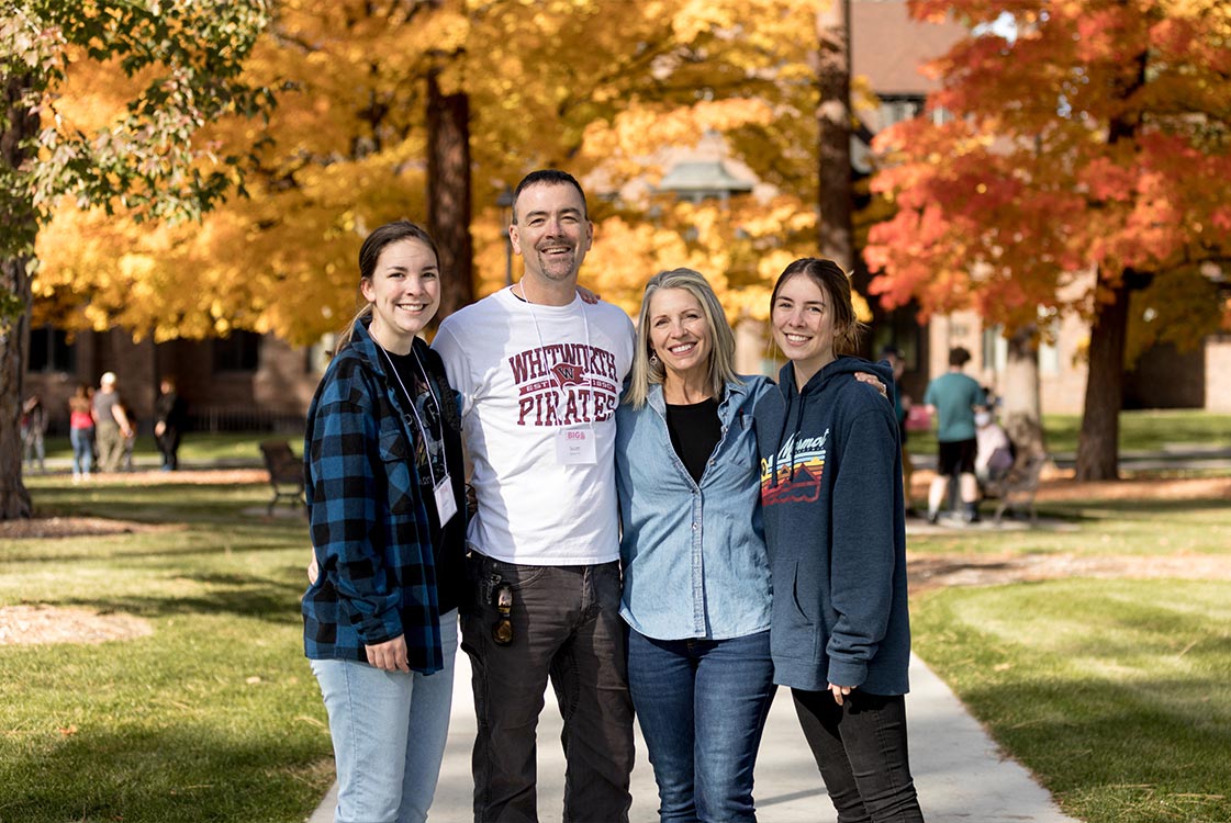Two parents and two students stand outside, smiling, with colorful autumn trees in the background.