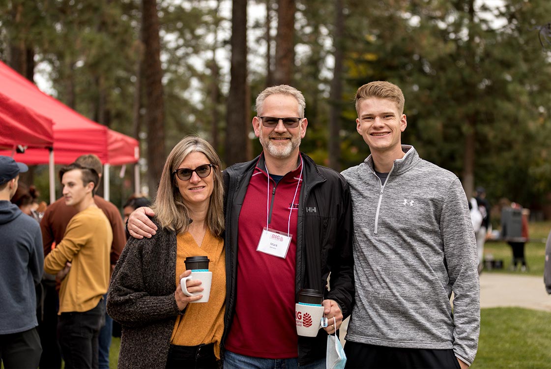 Two parents and a student pose together at an outdoor event, two of them holding coffee cups.