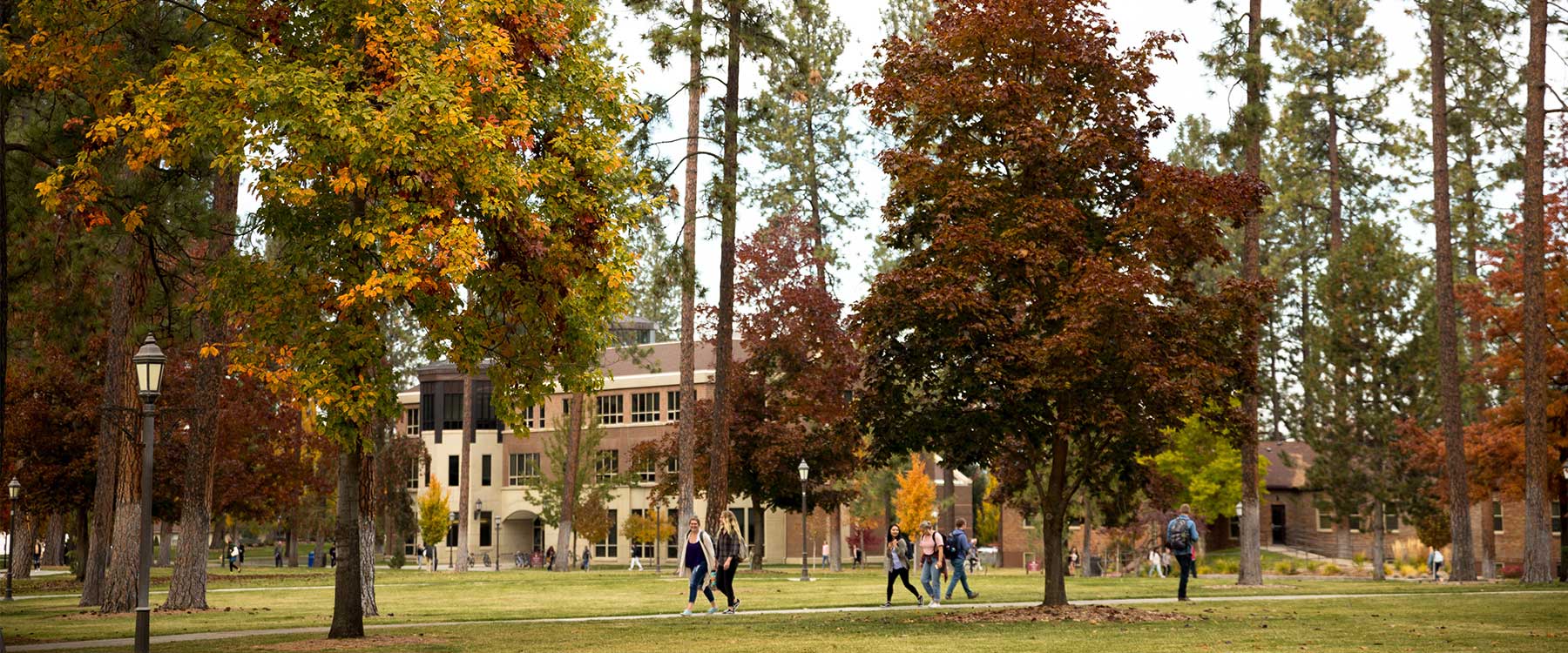 Many students walk between academic buildings on campus pathways beneath trees with fall foliage.