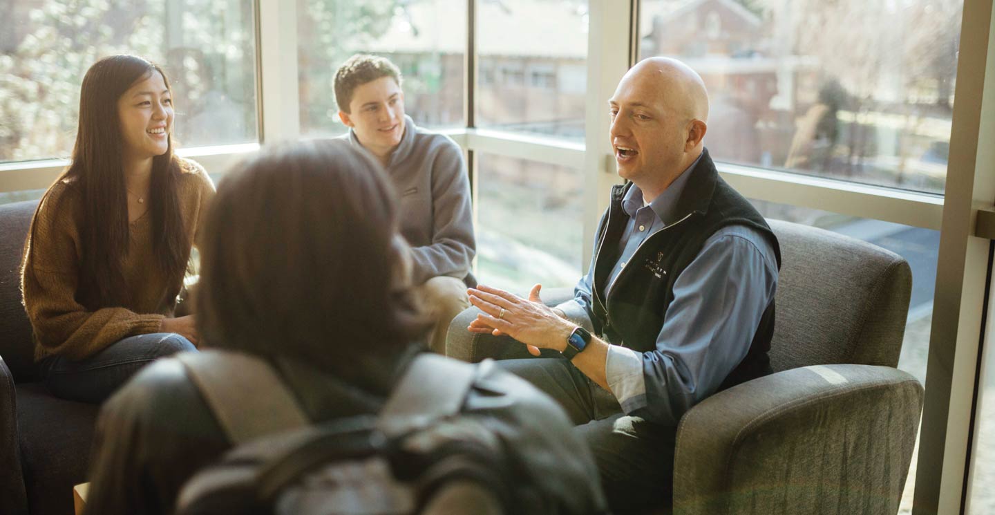 Three students and a professor engage in a discussion by the windows in a cozy, well-lit room.