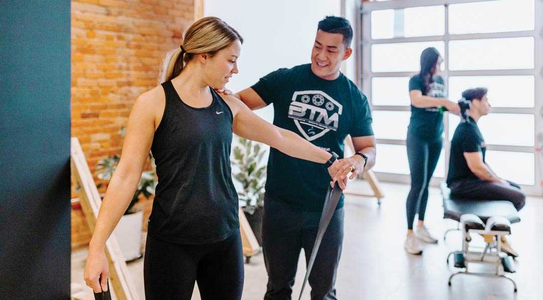 A fitness trainer helps a client with resistance bands while others exercise in the background.