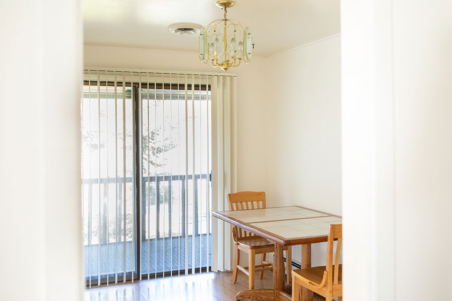 Kitchen nook with a wooden table, two chairs and sliding glass doors with vertical blinds.