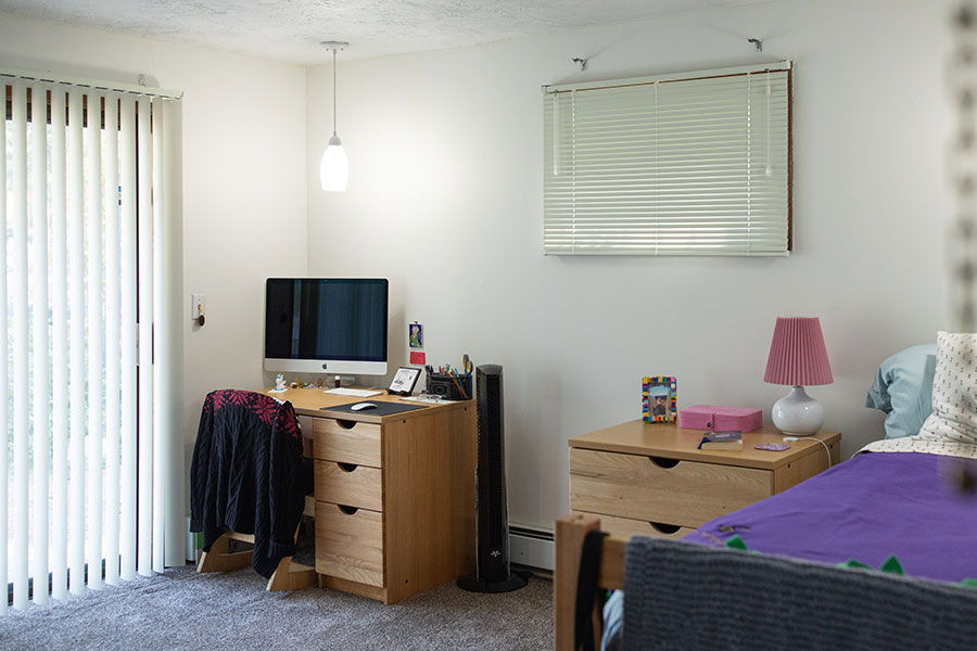 Bedroom with a computer desk, twin bed and bedside dresser. Blinds cover the window, and a sliding door lets in natural light.