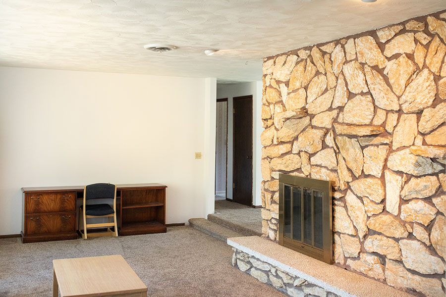 Living room with a large stone fireplace, beige carpet and a wooden desk.