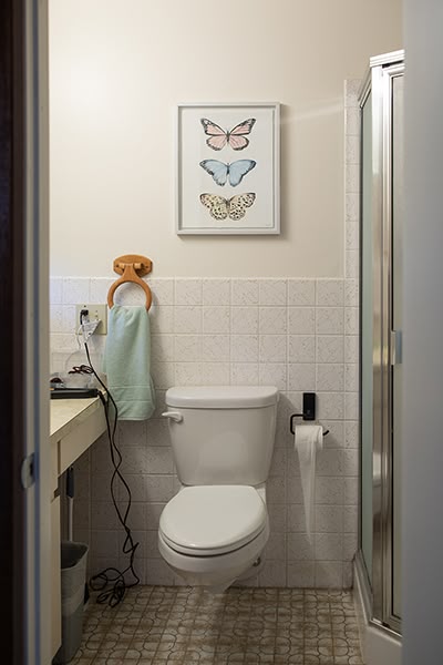 A tiled bathroom with a shower, toilet, sink, mirror and wall art.