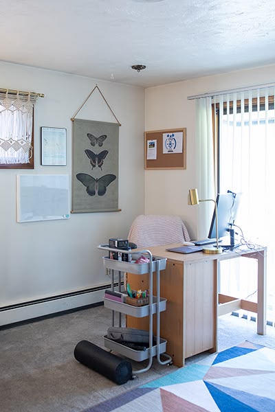 Corner of a bedroom set up as a home office with a desk, chair, computer and wall art. Natural light enters through a sliding door.