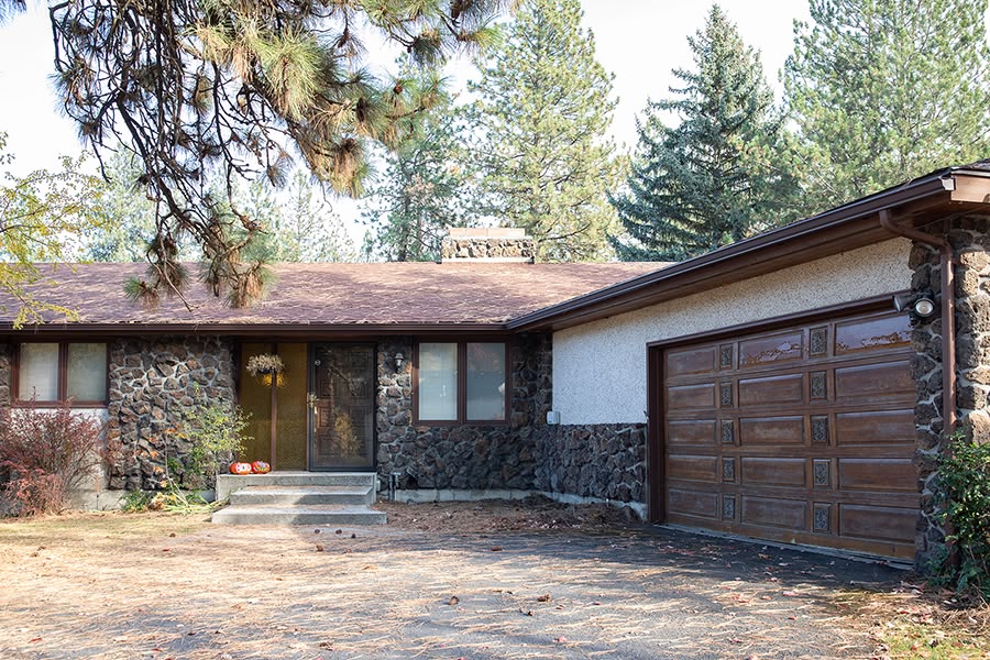 A rustic house with a stone facade, wooden garage door and pine trees in the background.