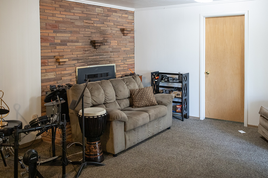 A cozy rec room with carpeted floors and a brick fireplace with insert. A sofa and drum set sit in front of the fireplace.