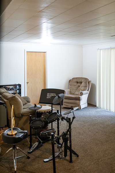 A cozy rec room with carpeted floors, a sofa and chair, and an electric drum set.